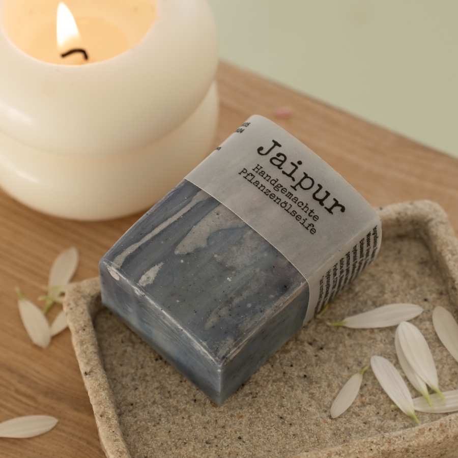 Handgemachte Jaipur Naturseife mit dunkelgrauer Marmorierung auf einer Naturstein-Seifenschale neben einer brennenden Kerze.